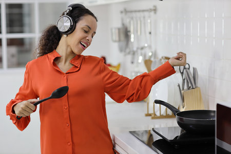 Happy latin woman cooking, smelling and tasting food in kitchen. Beautiful young female with headphone preparing delicious meal while listen music at home. Healthy and lifestyle conceptの写真素材