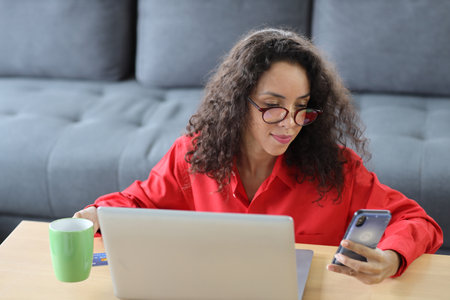 Happy woman sitting and using computer and holding credit card for online payment or shopping in living room. Businesswoman showing payment completed with raising hands and exciting emotion.の写真素材