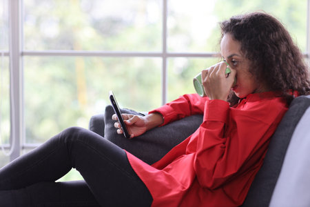 Happy smiling latin woman sitting on sofa while drinking coffee and using internet on smart mobile phone in living room. Lifestyle with technologyの写真素材