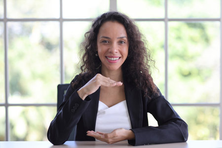 Attractive professional latin female in classic suit show protecting something on table. Businesswoman sitting at home workplace. Property insurance and security conceptの写真素材