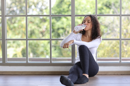 Charming African American holding and drinking bottle of water drink clean clear mineral aqua for healthy and body refreshment after finish yoga excercise. Happy female sitting in living room at homeの写真素材
