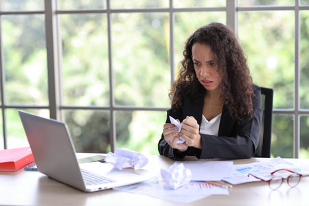 Mid adult businesswoman touching her head with crumpled paper on table in office after bad news...
