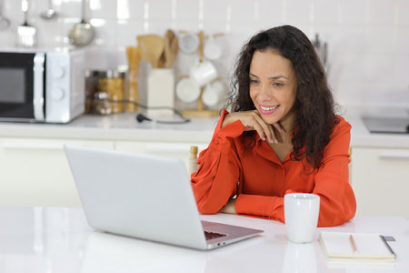 Smart and active latin woman sitting and using internet on computer while using smart mobile phone with a cup of coffee in kitchen. Anywhere and work. Lifestyle with technologyの写真素材