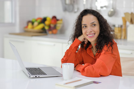 Smart and active latin woman sitting and using internet on computer while using smart mobile phone with a cup of coffee in kitchen. Anywhere and work. Lifestyle with technologyの写真素材