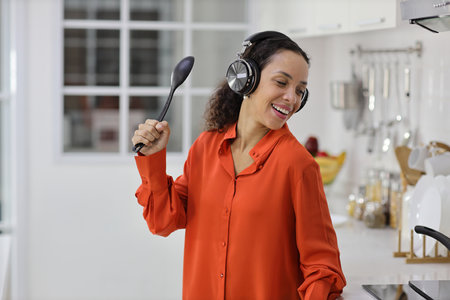 Happy latin woman cooking, smelling and tasting food while dancing in kitchen. Beautiful young female with headphone preparing delicious meal while listen music at home. Healthy and lifestyle conceptの写真素材