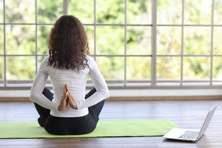 Sporty young adult woman sit on yoga mat practicing mediate yoga watching coach training video online from laptop. Healthy female doing exercise and taking lesson distant course among nature outdoorの写真素材