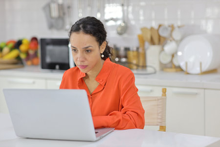 Smart and active latin woman sitting and using internet on computer while using smart mobile phone with a cup of coffee in kitchen. Anywhere and work. Lifestyle with technologyの写真素材