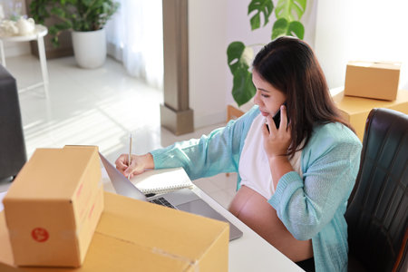 Attractive happy asian pregnant woman sitting and using mobile smart phone and checking order for customer with tablet. Expectant mother preparing and working for baby birth during pregnancy concept.の写真素材