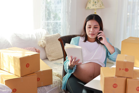 Attractive happy asian pregnant woman sitting and using mobile smart phone and checking order for customer with tablet. Expectant mother preparing and working for baby birth during pregnancy concept.の写真素材