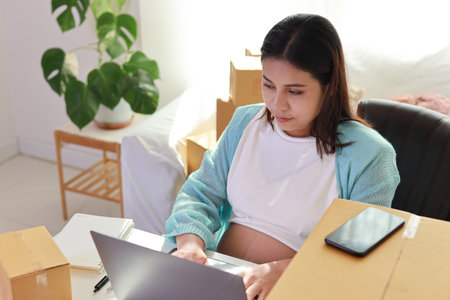 Attractive happy asian pregnant woman sitting and using mobile smart phone and making order for customer with computer. Expectant mother preparing and working for baby birth during pregnancy conceptの写真素材