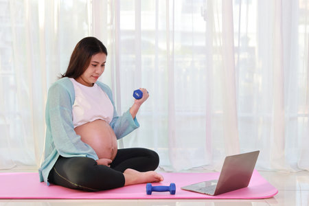 Happy young asian pregnant sit on pink mat lifting dumbbell and watch training video online from computer in living room at home. Expectant mother prepare for baby birth during pregnancy conceptの写真素材