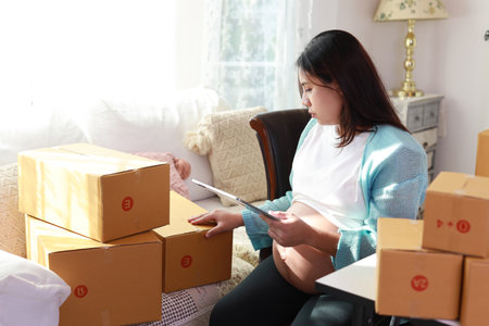 Attractive happy asian pregnant woman sitting and using mobile smart phone and writing order for customer with tablet. Expectant mother preparing and working for baby birth during pregnancy concept.の写真素材