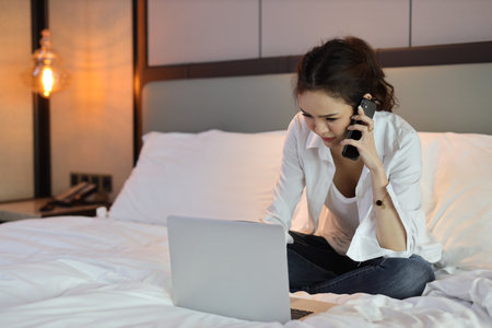 Active and unhappy asian woman in casual sitting on bed and working smart mobile phone and computer in hotel, beauty businesswoman with bedroom background. Lifestyle technology conceptの写真素材
