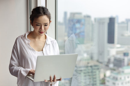 Portrait of confident and beautiful young asian businesswoman with happy emotion standing and using computer in modern hotel with city blurred background. Success business and lifestyle conceptの写真素材