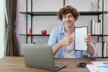 Caucasian businessman online meeting with colleage or customer and showing something on white screen tablet while sitting at home and showing thumb upの写真素材