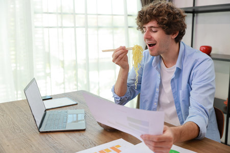 Young caucasian currly hair man sitting on table and eating tasty and instant noodle for lunch in the working room with chopsticks and computer. Unhealthy and cheap food lifestyle conceptncept.の写真素材