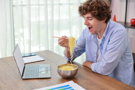 Young caucasian currly hair man sitting on table and eating tasty and instant noodle for lunch in the working room with chopsticks and computer. Unhealthy and cheap food lifestyle conceptncept.の写真素材