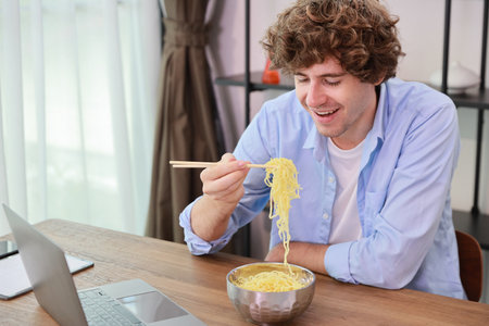 Young caucasian currly hair man sitting on table and eating tasty and instant noodle for lunch in the working room with chopsticks and computer. Unhealthy and cheap food lifestyle conceptncept.の写真素材