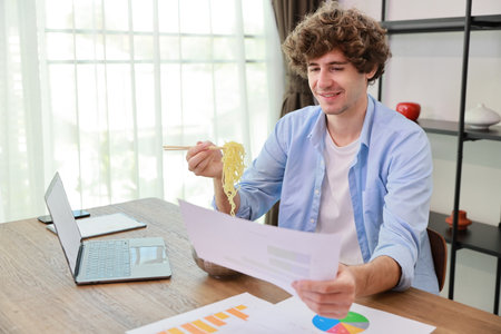 Young caucasian currly hair man sitting on table and eating tasty and instant noodle for lunch in the working room with chopsticks and computer. Unhealthy and cheap food lifestyle conceptncept.の写真素材