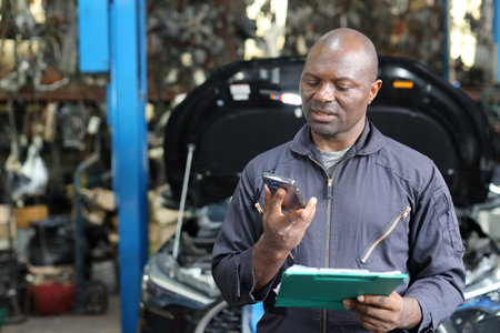 Man technician car mechanic in uniform checking maintenance a car service with tablet while using smart mobile phone at repair garage station. Car repair service conceptの写真素材