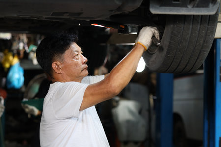 Man technician car mechanic in half uniform checking maintenance a lifted car service at repair garage station. Worker holding wrench and fixing breakdown vehicle. Concept of car center repair serviceの写真素材