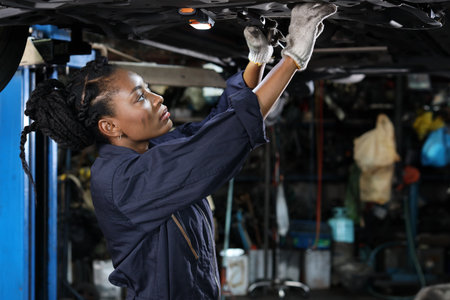 Woman technician car mechanic in uniform checking maintenance a lifted car service at repair garage station. Worker using flashlight and wrench fixing breakdown vehicle. Car repair service concept.の写真素材