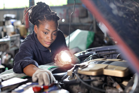 Woman technician car mechanic in uniform checking maintenance car service at repair garage station. Worker holding flashlight and wrench fixing breakdown vehicle. Concept of car center repair service.の写真素材