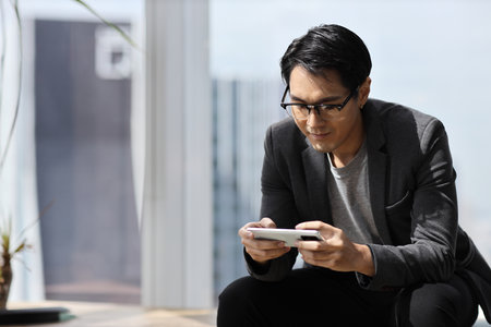 Young asian business man sitting and using smart mobile phone in business office building and city backgroundの写真素材