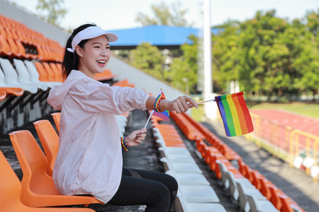 Happy and excited young female asian transgender LGBT sport fan cheering and watching the match with rainbow flag pointing and wristband, she sitting amongst the rows of empty seats on stadiumの写真素材