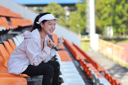 Portrait of happy and exciting young female asian sport fan cheering and watching the match with her hands fisting, she sitting amongst the rows of empty seats on stadium while celebrate winner teamの写真素材