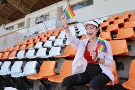 Happy and excited young female asian transgender LGBT sport fan cheering and watching the match with rainbow flag raising and wristband, she sitting amongst the rows of empty seats on stadiumの写真素材