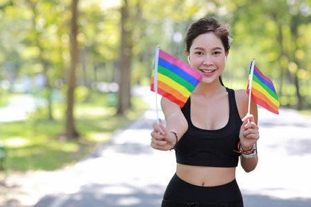 Young cheerful asian model transgender LGBT with wristband offering someone a rainbow flag while smiling and looking at camera. Photo of sporty woman runner in sportswear. pride and equality concept.の写真素材