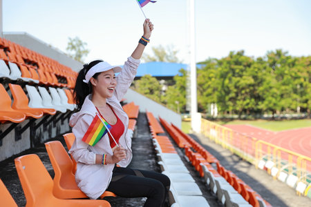 Happy and excited young female asian transgender LGBT sport fan cheering and watching the match with rainbow flag raising and wristband, she sitting amongst the rows of empty seats on stadiumの写真素材
