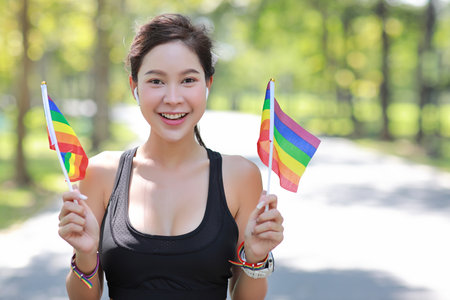 Young cheerful asian model transgender LGBT with wristband waving rainbow flag while smiling and looking at camera. Photo of attractive sporty woman runner in sportswear. pride and equality concept.の写真素材