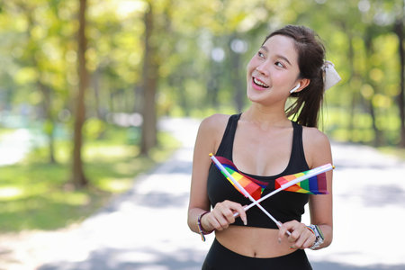Young cheerful asian model transgender LGBT with wristband waving rainbow flag while smiling and looking at camera. Photo of attractive sporty woman runner in sportswear. pride and equality concept.の写真素材