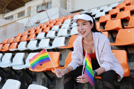Happy and excited young female asian transgender LGBT sport fan cheering and watching the match with rainbow flag raising and wristband, she sitting amongst the rows of empty seats on stadiumの写真素材
