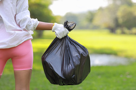 Rear view young beautiful asian volunteer woman hands hold garbage bag and picking up litter while cleaning plastic bottle from lawn during a volunteering environmental cleanup area park event outdoorの写真素材