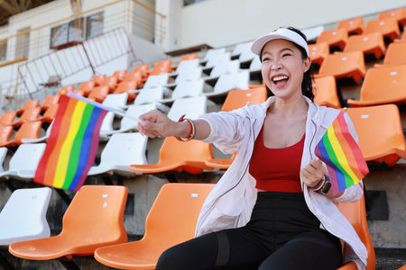 Happy and excited young female asian transgender LGBT sport fan cheering and watching the match with rainbow flag raising and wristband, she sitting amongst the rows of empty seats on stadiumの写真素材