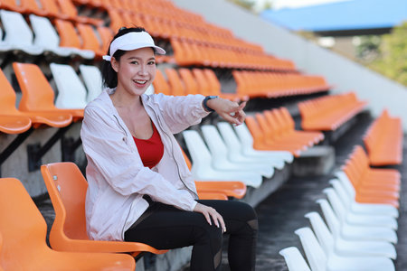 Portrait of happy and excited young female asian sport fan cheering and watching the match with her finger pointing, she sitting amongst the rows of empty seats on stadium while celebrate winner teamの写真素材