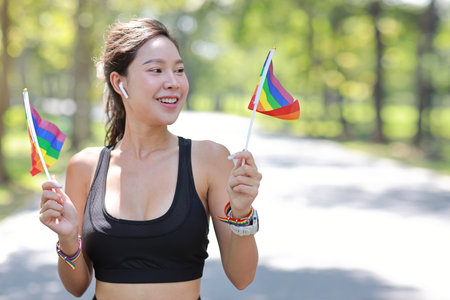 Young cheerful asian model transgender LGBT with wristband waving rainbow flag while smiling and looking at camera. Photo of attractive sporty woman runner in sportswear. pride and equality concept.の写真素材