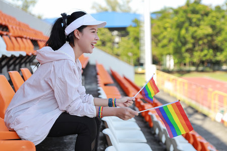 Happy and excited young female asian transgender LGBT sport fan cheering and watching the match with rainbow flag pointing and wristband, she sitting amongst the rows of empty seats on stadiumの写真素材