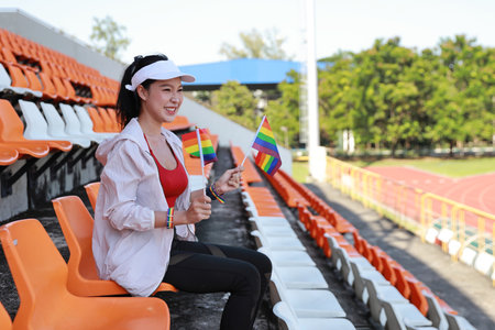 Happy and excited young female asian transgender LGBT sport fan cheering and watching the match with rainbow flag pointing and wristband, she sitting amongst the rows of empty seats on stadiumの写真素材