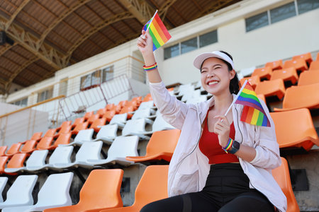 Happy and excited young female asian transgender LGBT sport fan cheering and watching the match with rainbow flag raising and wristband, she sitting amongst the rows of empty seats on stadiumの写真素材