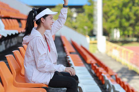 Portrait of happy and excited young female asian sport fan cheering and watching the match with her hands clapping, she sitting amongst the rows of empty seats on stadium while celebrate winner teamの写真素材