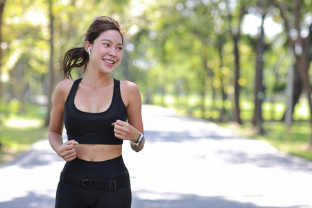 Beautiful asian fitness female sportswoman runner doing morning jogging exercise training outdoor. Happy smile athlete woman running under the tree in the park while looking at camera and listen musicの写真素材