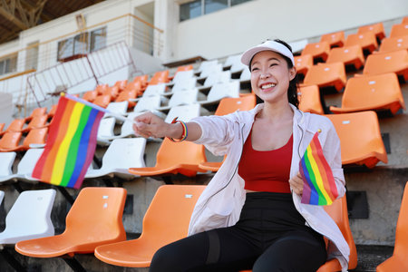Happy and excited young female asian transgender LGBT sport fan cheering and watching the match with rainbow flag pointing and wristband, she sitting amongst the rows of empty seats on stadiumの写真素材