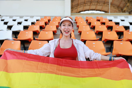 Happy and excited young female asian transgender LGBT sport fan cheering and watching the match with rainbow flag raising and wristband, she sitting amongst the rows of empty seats on stadiumの写真素材