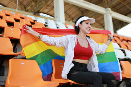 Happy and excited young female asian transgender LGBT sport fan cheering and watching the match with rainbow flag raising and wristband, she sitting amongst the rows of empty seats on stadiumの写真素材