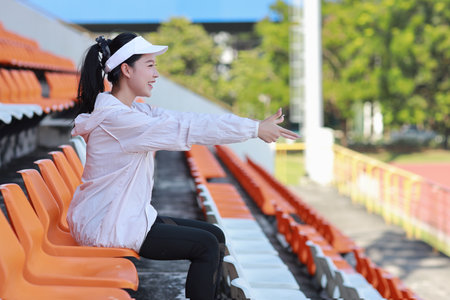 Portrait of happy and excited young female asian sport fan cheering and watching the match with her finger pointing, she sitting amongst the rows of empty seats on stadium while celebrate winner teamの写真素材