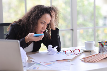 Mid adult businesswoman touching her head with crumpled paper on table in office after bad news business failure or get fired and feeling discouraged, distraught and hopeless in modern office.の写真素材